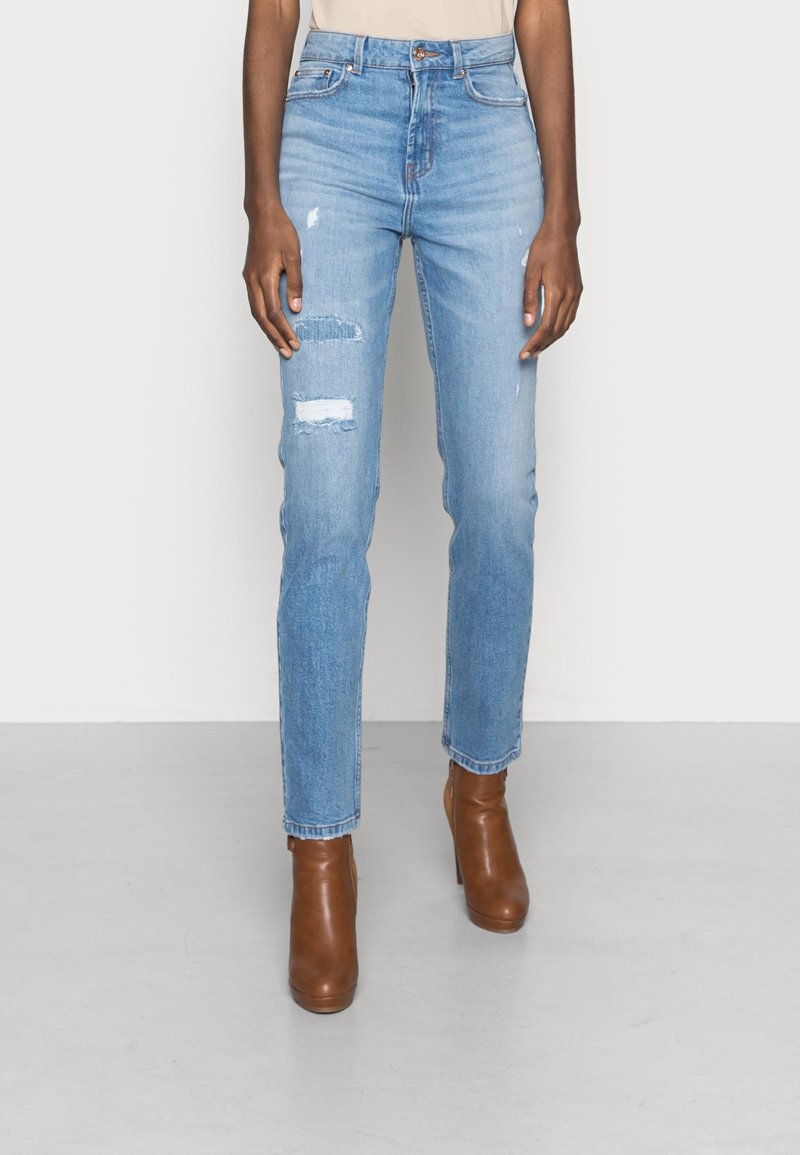 Jean slim bleu clair avec des déchirures réparées sur la cuisse gauche, porté avec des bottines marron à talons.
