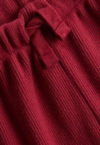 Roter, gerippter Stoff mit einer weichen Textur, der über eine Kordelzugdetail zur Anpassung verfügt. Das Material wirkt warm und gemütlich.