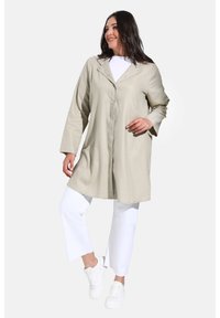 Veste longue beige clair en tissu texturé, avec un devant à boutons, des manches 3/4 et une coupe décontractée, associée à un pantalon et des chaussures blancs.