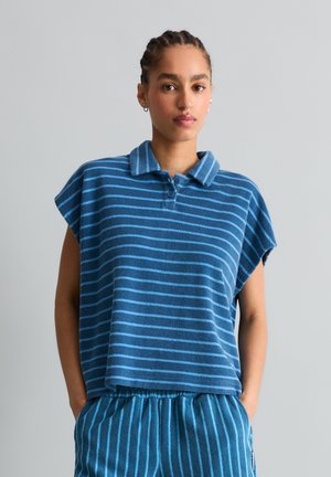 Jong persoon met gevlochten haar draagt een blauw kraaghemd met korte mouwen en bijpassende gestreepte broek, staand tegen een effen grijze achtergrond.