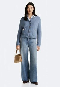 Mulher vestindo um cardigã azul com botões, camisa branca, jeans de perna larga, botas castanhas, segurando uma pequena bolsa castanha.