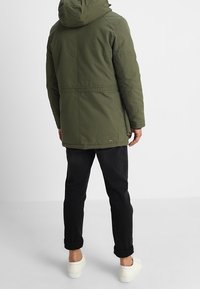 Chaqueta verde con capucha, cierre de cremallera, puños elásticos, cintura con cordón ajustable y un sutil detalle de logo. Combinada con pantalones negros enrollados y zapatos blancos.