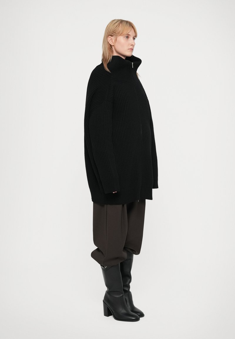 Chaqueta de punto negra de ribete grueso y corte holgado con cuello alto, cierre de cremallera y hombros caídos, combinada con pantalones marrones holgados y botas negras de tacón.