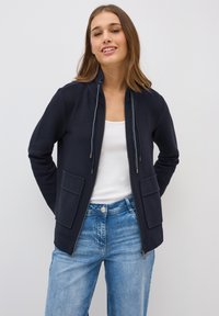 Navy Zip-up-Jacke mit zwei Fronttaschen, Kordelzugkapuze und gerippter Textur. Über einem weißen Shirt getragen, kombiniert mit blauen Jeans.