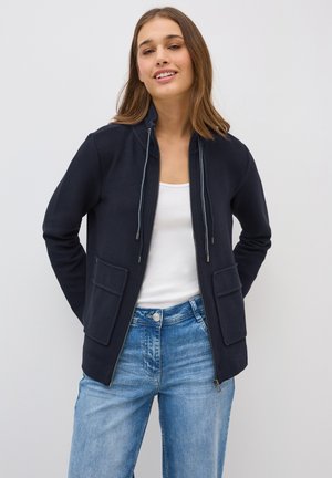 Veste zippée marine avec deux poches avant, capuche avec cordon de serrage et texture côtelée. Portée par-dessus une chemise blanche, associée à un jean en denim bleu.