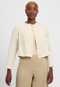 Light beige cropped jacket with long sleeves, round neck, front button closure, and two patch pockets. Made from textured, lightweight fabric.