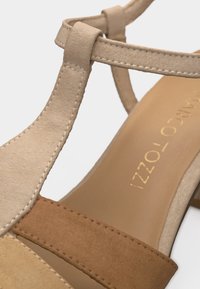 Γυναικεία σανδάλια από μπεζ και καφέ σαουέτ με σχέδιο T-λαιμού και ορατή ραφή στους ιμάντες και στη σόλα.