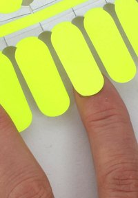 Hand mit Fingern, die leuchtend gelbe selbstklebende Nagelstreifen auf eine helle Oberfläche auftragen, Nahaufnahme zeigt teilweise Abdeckung der Nägel.