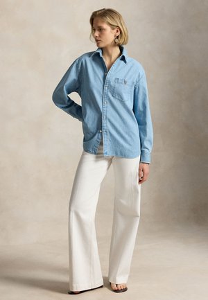 Kvinde med kort blondt hår iført en lyseblå denimskjorte, hvide vidde bukser og sorte sandaler, stående mod en beige baggrund.