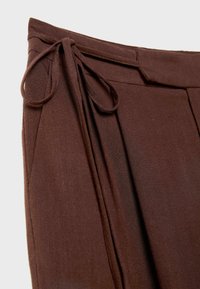 Bruna, veckade byxor med ett texturerat tyg. Har dragsko i midjan och sidofickor för extra funktionalitet.