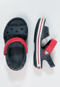 Sandales noires et rouges avec sangle réglable, trous de ventilation, semelle intérieure texturée et détail de semelle rayée blanche. Matériau léger et durable.