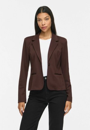 Femme aux longs cheveux foncés portant un blazer marron foncé, un haut blanc et un pantalon noir, debout devant un fond clair uni.
