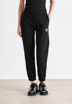 Osoba ubrana w czarne zwężane spodnie dresowe z elastycznymi ściągaczami i białym logo przy kieszeni, w połączeniu z czarno-białymi trampkami.