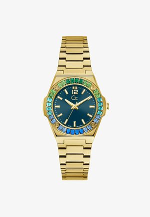 Kuldtooni kell, millel on ristkülikukujuline korpus, sinine dials ja kristallbezel rohelise ja sinise varjundiga. Metallsideme rihm läikiva viimistlusega.