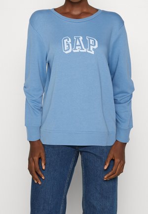 Person trägt hellblauen langärmligen GAP-Sweatshirt und blaue Jeans, steht mit entspannten Armen an den Seiten vor einem schlichten Hintergrund.