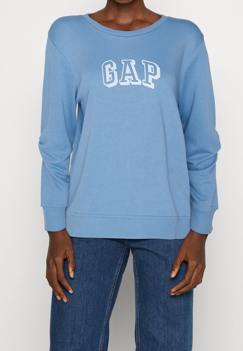 Personne portant un sweat-shirt GAP à manches longues bleu clair et un jean bleu, debout avec les bras détendus le long du corps contre un fond uni.