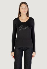 Maglietta nera a maniche lunghe con scollatura rotonda e logo metallico "Guess" decorato con strass sul davanti. Abbinata a jeans scuri.