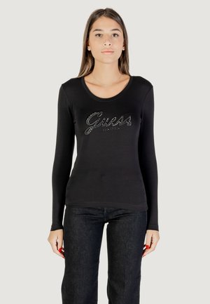 Maglietta nera a maniche lunghe con scollatura rotonda e logo metallico "Guess" decorato con strass sul davanti. Abbinata a jeans scuri.