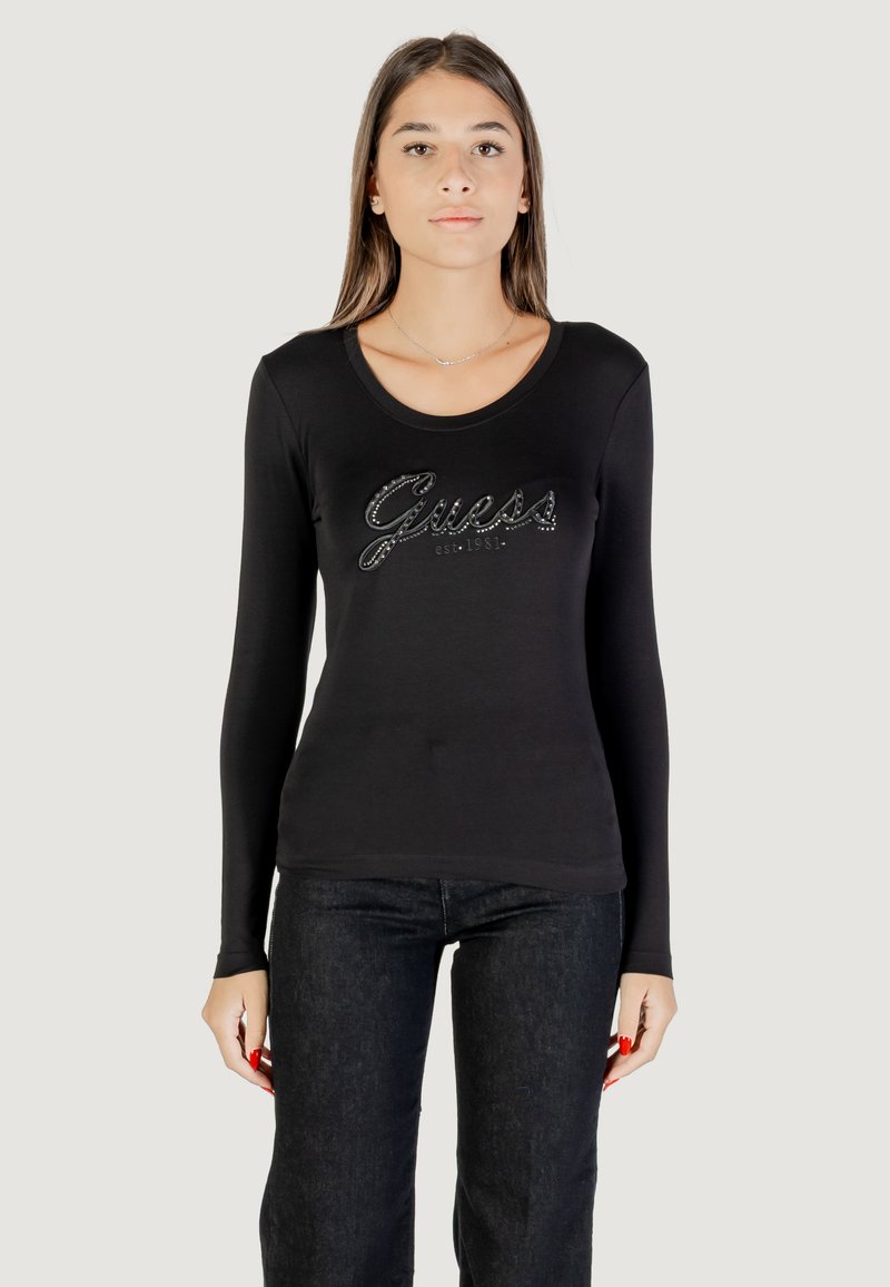 Maglietta nera a maniche lunghe con scollatura rotonda e logo metallico "Guess" decorato con strass sul davanti. Abbinata a jeans scuri.