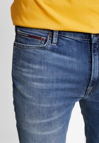 Modré džíny z denimového materiálu s vybledlým designem, obsahující pět kapes a viditelný červený akcent v prošívání přední kapsy.