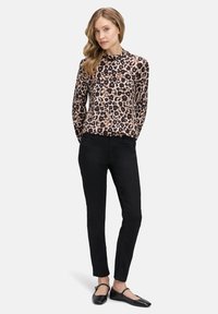 Dlhý rukáv leopardí potlač top v béžovej a hnedej, spárovaný s čiernymi slim-fit nohavicami a čiernymi plochými topánkami. Modelka stojí s uvoľnenými rukami.