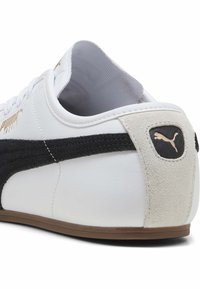 Baskets en cuir blanc avec une bande en suède noir, un logo doré et un onglet de talon en suède beige. Elles présentent un design traditionnel à lacets et une semelle en caoutchouc.