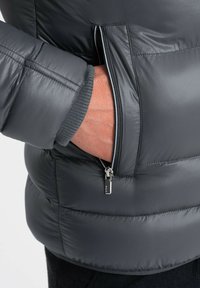Ręka w częściowo zapiętej bocznej kieszeni szarej ocieplanej kurtki noszonej na ciemnym mankiecie.
