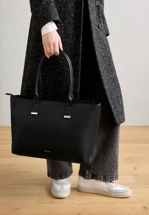 Person in white sneakers, gray pants, and dark textured coat holding a large black leather tote bag with gold accents.