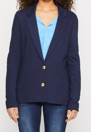 Nainen, jolla on yllään tummansininen bleiseri kultaisilla napeilla vaaleansinisen paidan ja tummansinisten farkkujen päällä, seisoo yksinkertaisen taustan edessä.