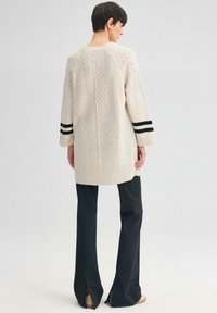 Beige gebreide trui met V-patroon, driekwartmouwen met zwarte strepen, gecombineerd met high-waisted zwarte broek met gespleten zomen.