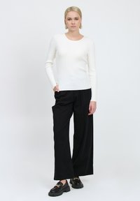 Top bianco a maniche lunghe con righe, abbinato a pantaloni neri larghi. Scarpe nere con un dettaglio a catena. Design semplice su uno sfondo neutro.