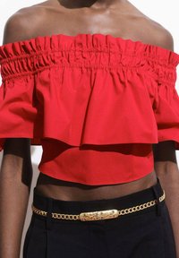 Top rosso senza spalle con strati arricciati, realizzato in tessuto leggero; abbinato a pantaloni neri a vita alta e una cintura in catena d'oro.