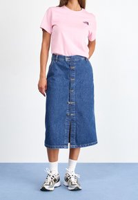 T-shirt rosa chiaro con logo, abbinato a una gonna midi in jeans blu a vita alta con bottoni davanti e orlo posteriore. Indossato con calzini bianchi e sneakers.