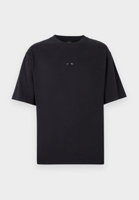 T-shirt noir à manches courtes avec un petit logo ovale centré sur la poitrine, présenté sur un fond gris clair uni.