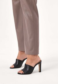 Mule din piele neagră, cu vârf deschis, cu o textură netedă, toc înalt tip stiletto și un design minimalist. Asortate cu pantaloni ajustați în nuanță taupe.
