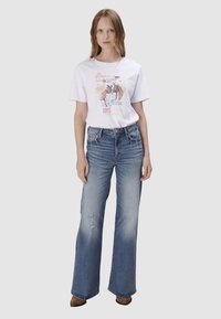 Witte grafische T-shirt met een kleurrijk rodeo-ontwerp, gecombineerd met lichtblauwe flare jeans met beschadigde details en bruine schoenen.