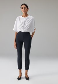 Witte top met korte mouwen en een gedraaide voorkant, gecombineerd met nauwsluitende, zwarte broek tot aan de enkels met openingen bij de manchetten en elegante zwarte mules.