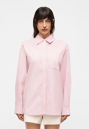 Γυναίκα με σκούρα μαλλιά φοράει ανοιχτό ροζ πουκάμισο με μακριά μανίκια και γιακά με διακριτικές κάθετες ρίγες και ανοιχτόχρωμο σορτς μπροστά σε απλό φόντο.