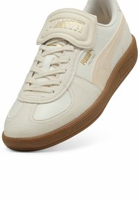 Bílé kožené tenisky s texturovaným povrchem, vyznačující se polstrovaným páskem a zlatými logovými akcenty, a hnědou gumovou podrážkou.