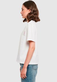 Hvid bomulds T-shirt med korte ærmer, en afslappet pasform og et lille pink broderet design på brystet. Ensfarvet baggrund.