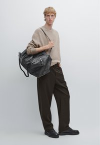 Svart läderväska med dragkedja, buren av en person i en beige tröja och mörka byxor, mot en neutral bakgrund.