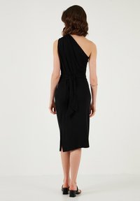 Black, one-shoulder dress with a tied waist, mid-calf length, and side slit. Made of smooth fabric with a sleek silhouette.