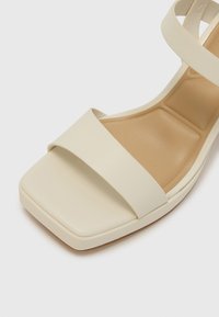 Sandale à talons en cuir crème avec un bout carré, de larges brides croisées, un dessus texturé et une base légèrement plateforme.