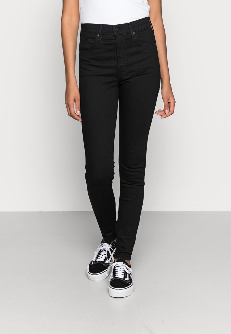 Jeans skinny negros de denim con una textura suave, de cintura alta, diseño de cinco bolsillos y cierre de botón. Llevados con zapatillas negras.
