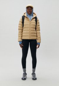 Chaqueta de plumas en beige claro, combinada con una capa base azul, leggings negros y zapatos de senderismo grises. Incluye una mochila y una gorra.