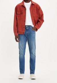 Röd denimjacka över en vit t-shirt, matchad med blå raka jeans och vita sneakers. Har två bröstfickor och knäppning med knappar.