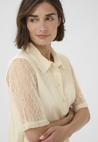 Frau mit kurzen braunen Haaren, die eine cremefarbene Spitzenbluse mit transparenten Ärmeln trägt und mit übergeschlagenen Armen nach links schaut.