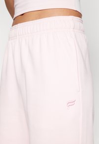 Light pink sweatpants featuring an elastic waistband, side pockets, and a small embroidered logo on the thigh. Material appears soft and smooth.
