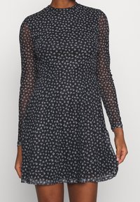 Schwarzes Blumenkleid mit langen, transparenten Ärmeln und hohem Halsauschnitt. Verfügt über einen schwingenden Rock und kleine weiße Blumenmuster auf dem gesamten Stoff.