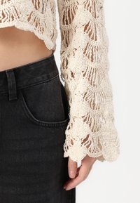 Top de crochê creme com mangas longas e largas, exibindo padrões em formato de escalope; combinado com jeans de denim preto ajustados, com um bolso frontal.